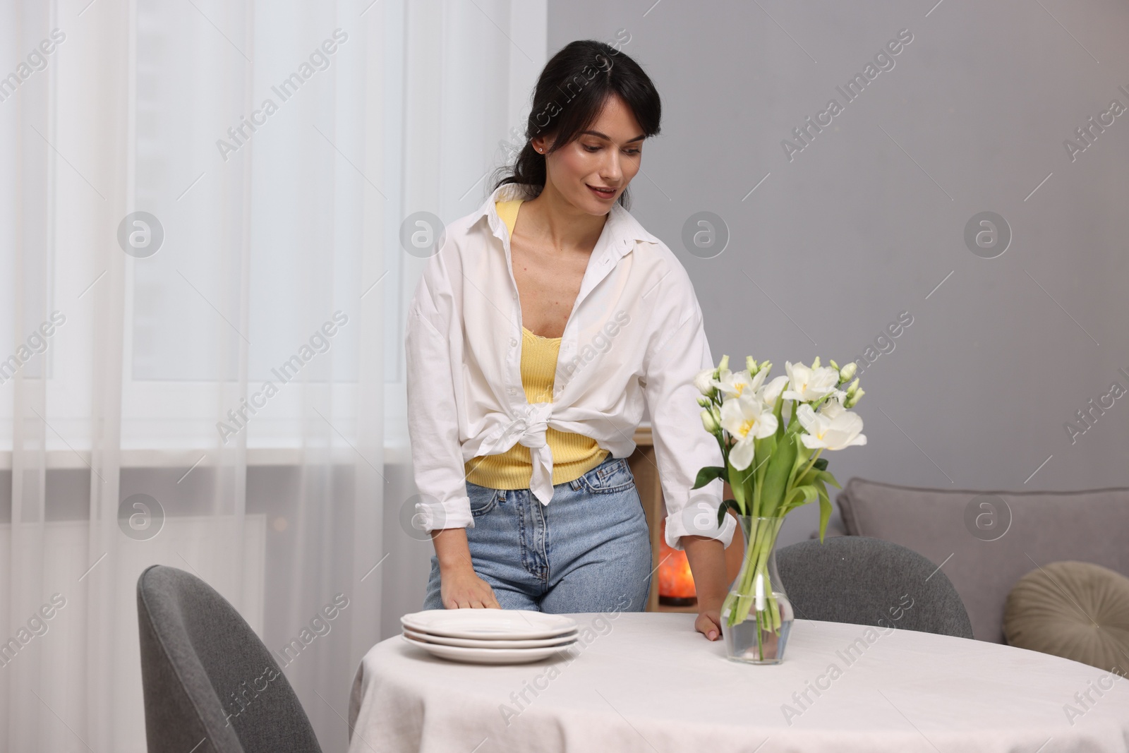 Photo of Young woman putting vase with flowers on table with white tablecloth at home