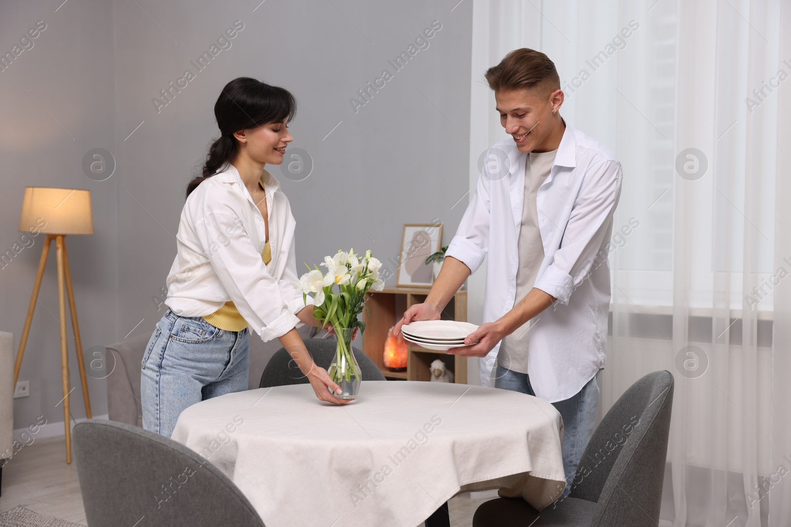 Photo of Couple putting plates and vase with flowers on table with white tablecloth at home