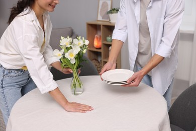 Photo of Couple putting plates and vase with flowers on table with white tablecloth at home, closeup