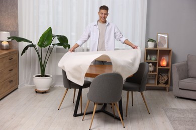 Photo of Young man putting white tablecloth on table at home