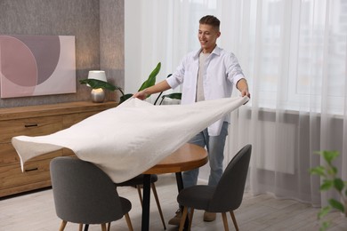 Photo of Young man putting white tablecloth on table at home