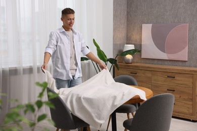 Photo of Young man putting white tablecloth on table at home