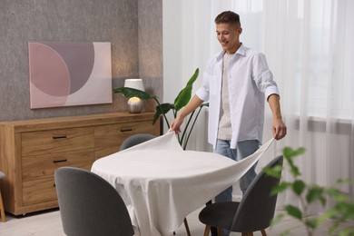 Young man putting white tablecloth on table at home