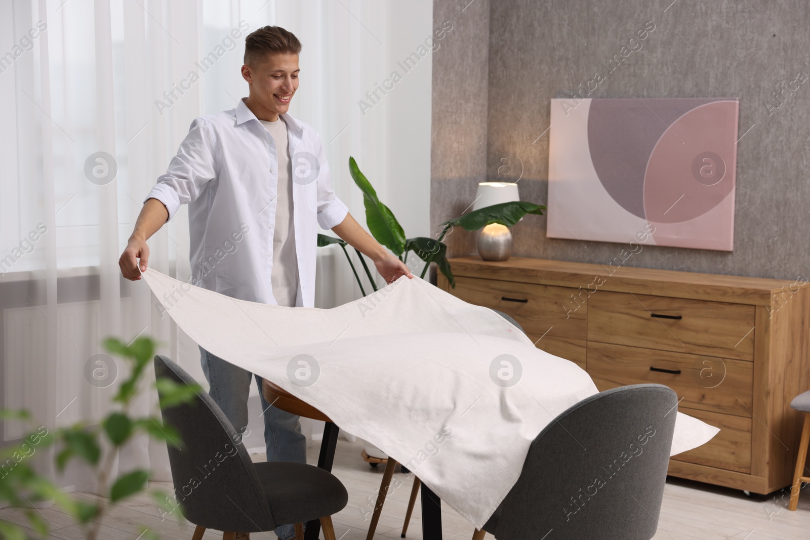 Photo of Young man putting white tablecloth on table at home