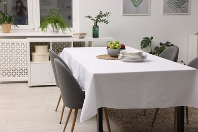 Photo of Table with white tablecloth, apples, plates and chairs in stylish room. Interior design