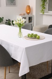 Photo of Table with white tablecloth, apples, flowers in vase and chairs in stylish room. Interior design