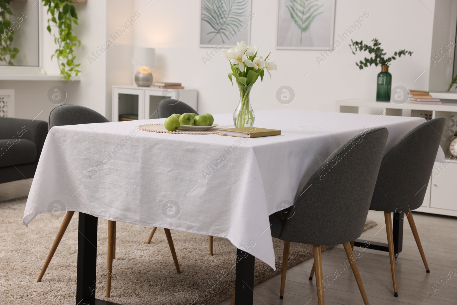 Photo of Table with white tablecloth, apples, flowers in vase and chairs in stylish room. Interior design