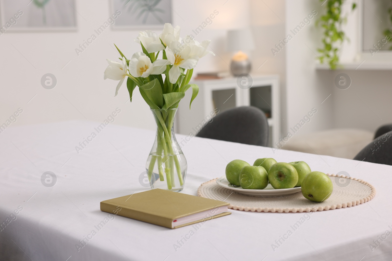 Photo of Apples, book and flowers in vase on table with white tablecloth indoors