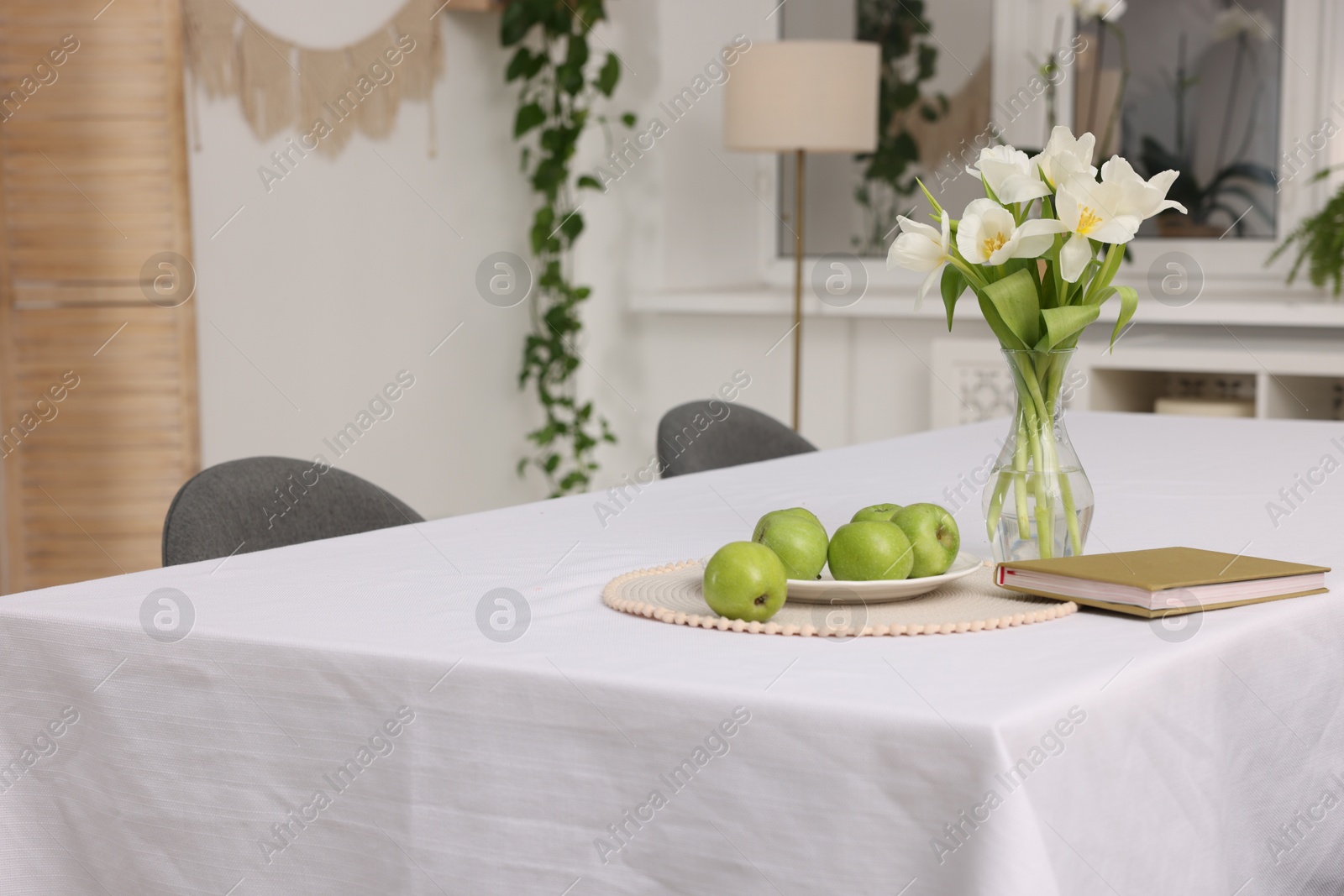 Photo of Apples, book and flowers in vase on table with white tablecloth indoors, space for text