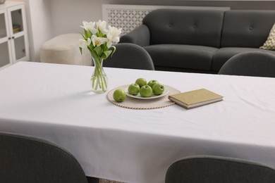 Photo of Apples, book and flowers in vase on table with white tablecloth indoors