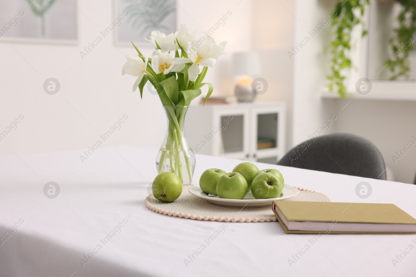 Photo of Apples, book and flowers in vase on table with white tablecloth indoors