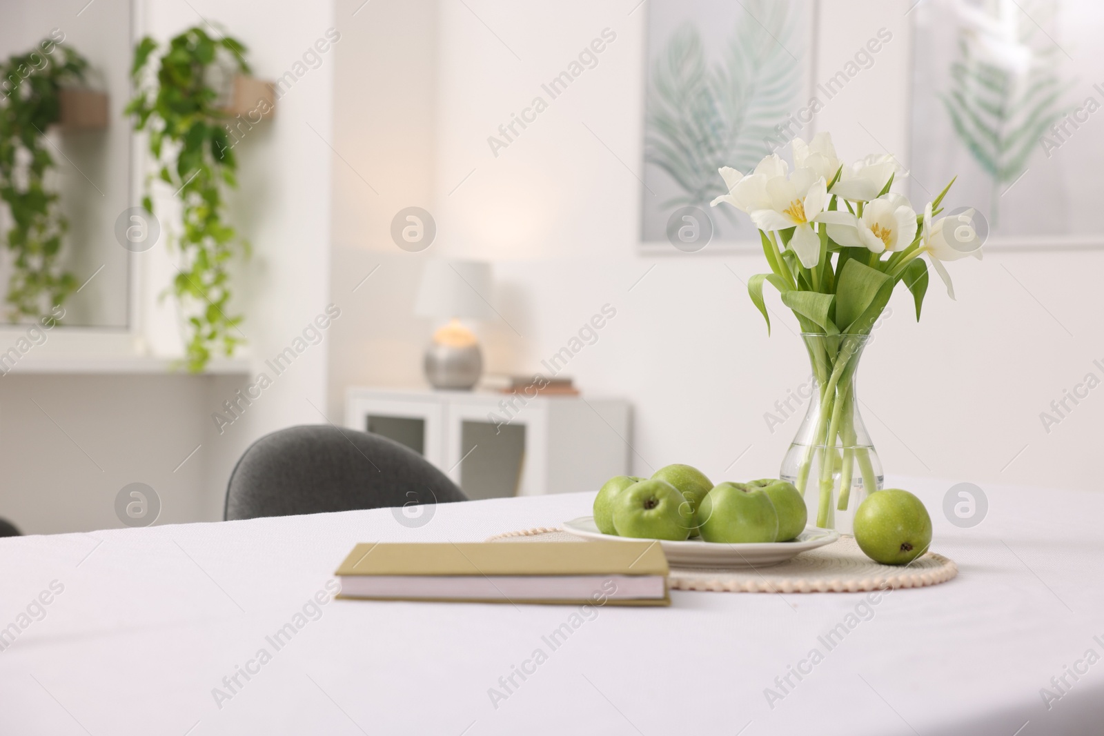 Photo of Apples, book and flowers in vase on table with white tablecloth indoors