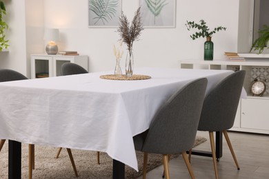 Photo of Table with white tablecloth, dried flowers in vases and chairs in stylish room. Interior design