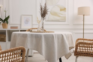 Photo of Table with white tablecloth, dried flowers in vases and chairs in stylish room. Interior design