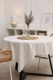 Photo of Table with white tablecloth, dried flowers in vases and chairs in stylish room. Interior design