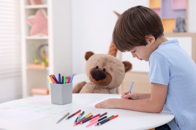 Photo of Cute boy drawing near teddy bear at white table in kindergarten. Space for text