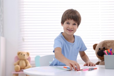 Photo of Smiling boy drawing near teddy bear at white table in kindergarten. Space for text