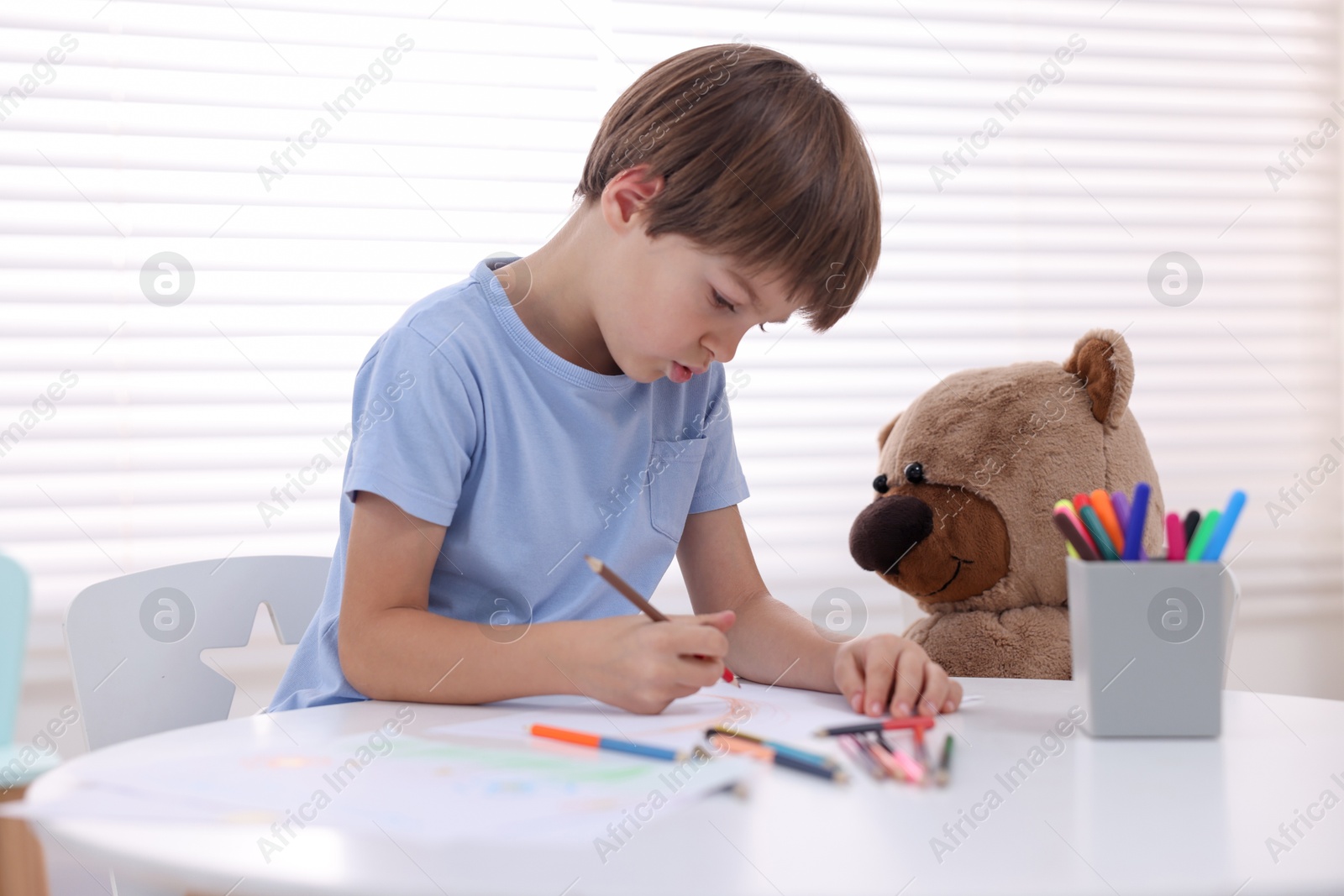 Photo of Cute boy drawing near teddy bear at white table in kindergarten