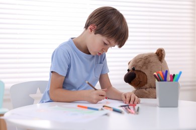 Photo of Cute boy drawing near teddy bear at white table in kindergarten