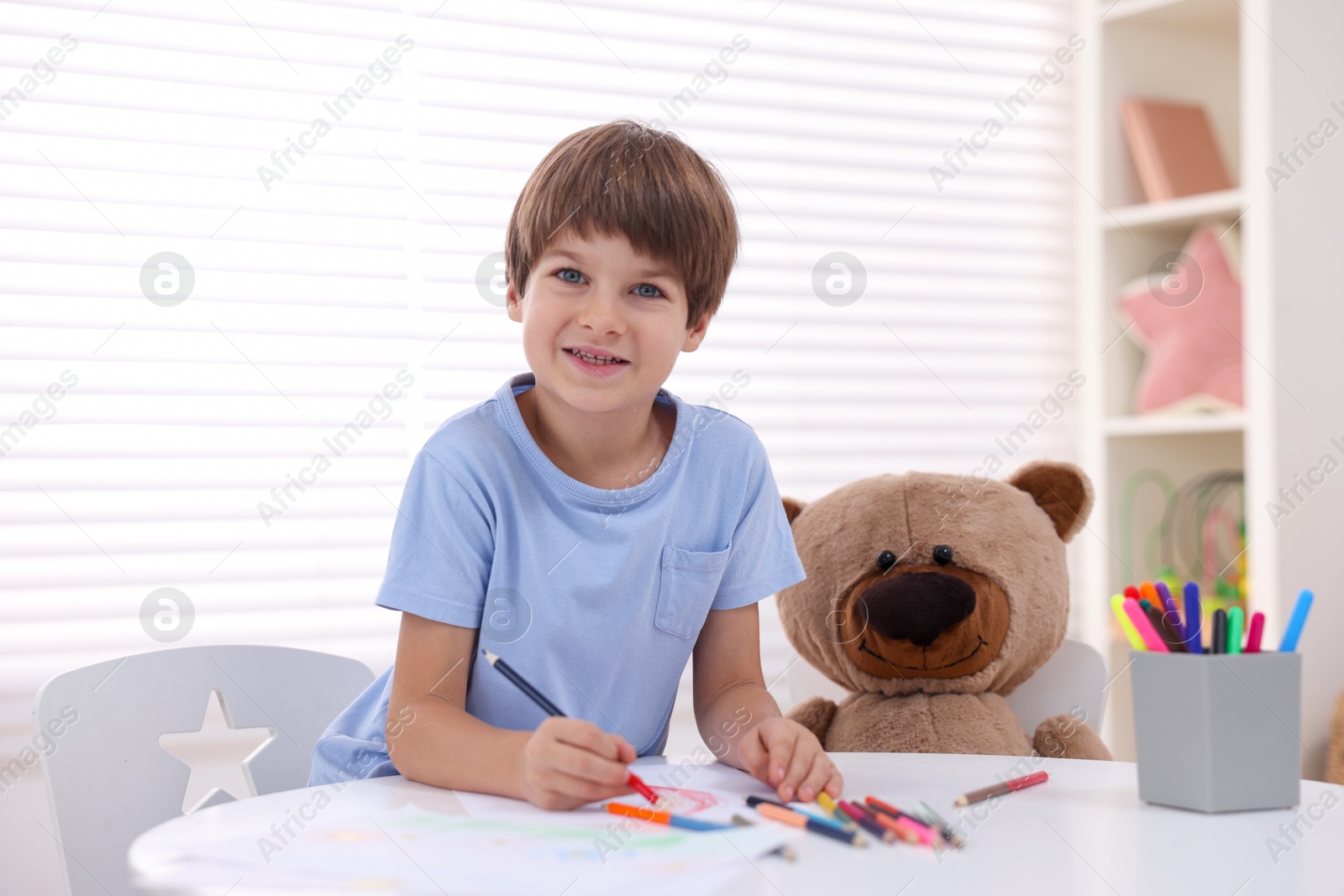 Photo of Smiling boy drawing near teddy bear at white table in kindergarten