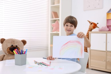 Photo of Smiling boy showing drawing at white table in kindergarten