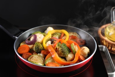 Photo of Frying pan with vegetables and stove on table against black background, closeup