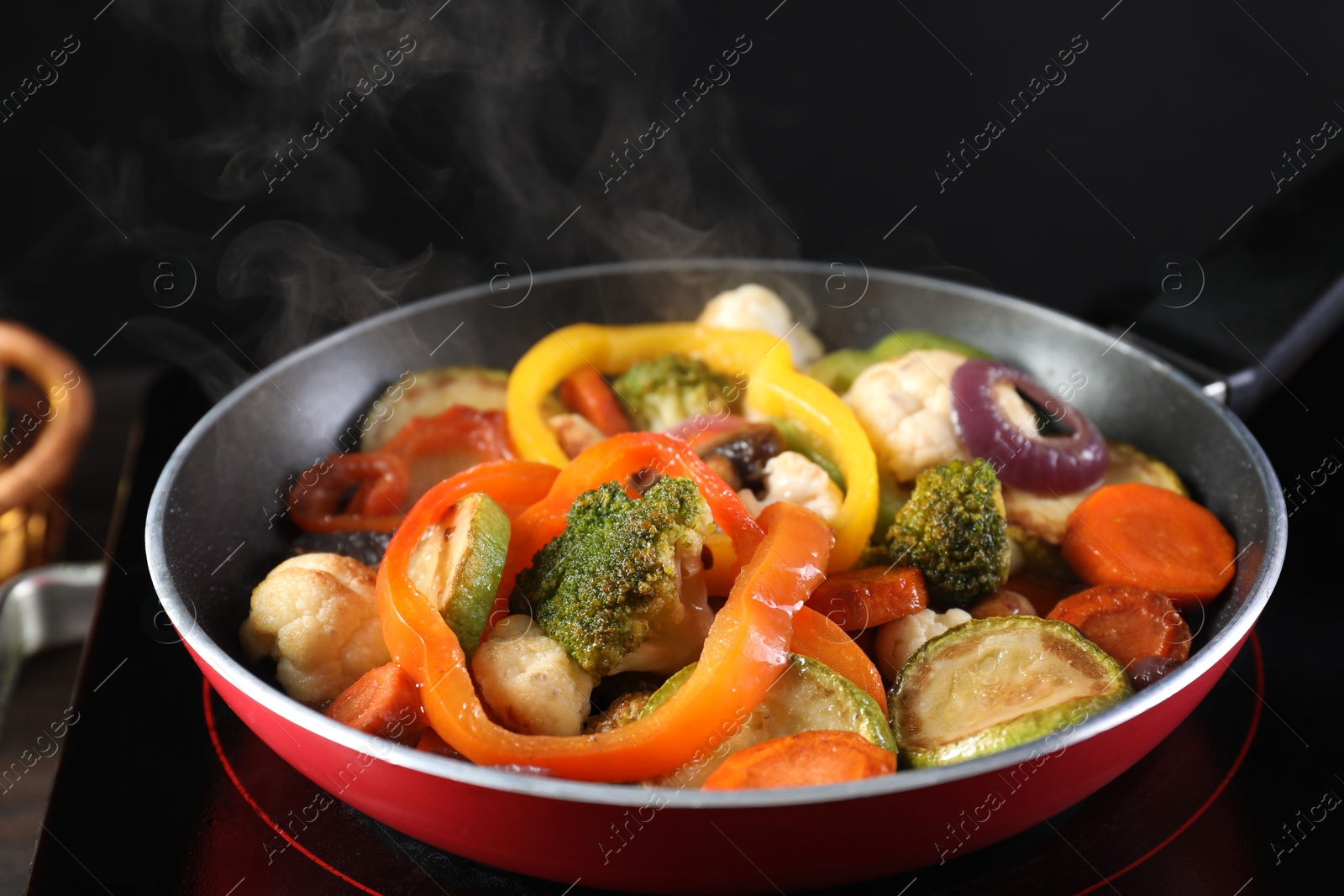 Photo of Frying pan with vegetables and stove on table against black background, closeup