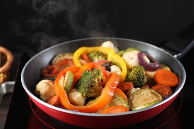 Photo of Frying pan with vegetables and stove on table against black background, closeup