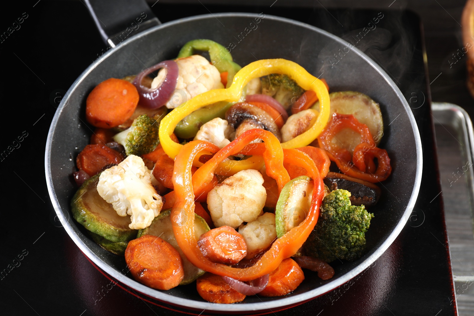 Photo of Frying pan with vegetables and stove on table against black background, closeup