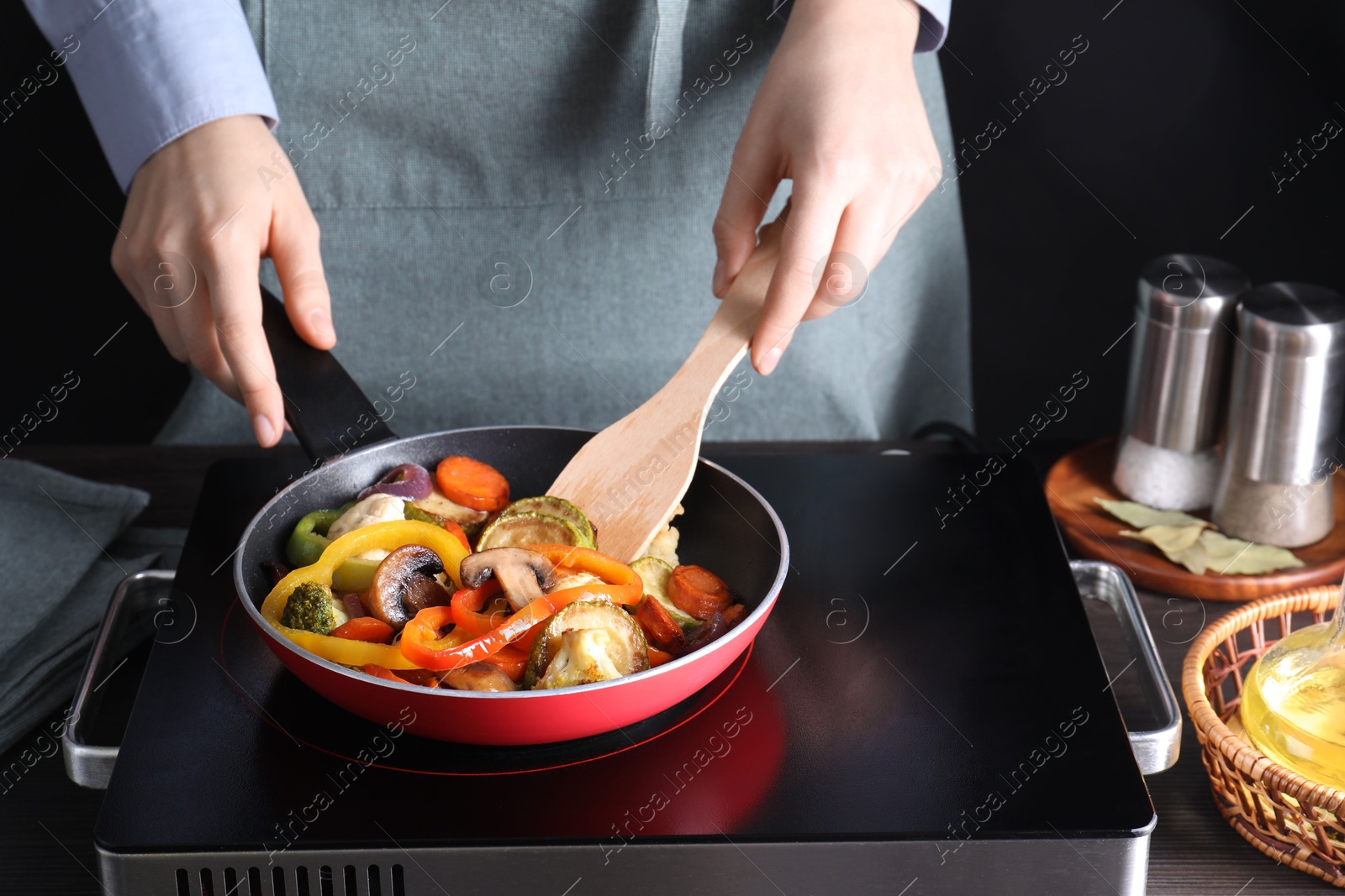 Photo of Woman frying vegetables and mushrooms in pan on stove at wooden table, closeup