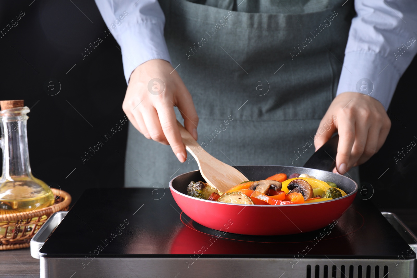 Photo of Woman frying vegetables and mushrooms in pan on stove at wooden table, closeup