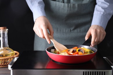 Photo of Woman frying vegetables and mushrooms in pan on stove at wooden table, closeup