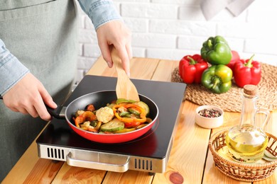 Photo of Woman frying vegetables in pan on stove at wooden table indoors, closeup