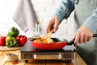 Photo of Woman frying vegetables in pan on stove at wooden table indoors, closeup