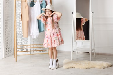 Photo of Stylish little girl wearing oversized high heeled shoes indoors