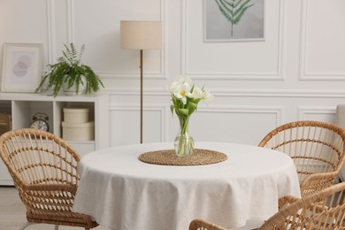 Photo of Table with white tablecloth, flowers in vase and chairs in stylish room. Interior design