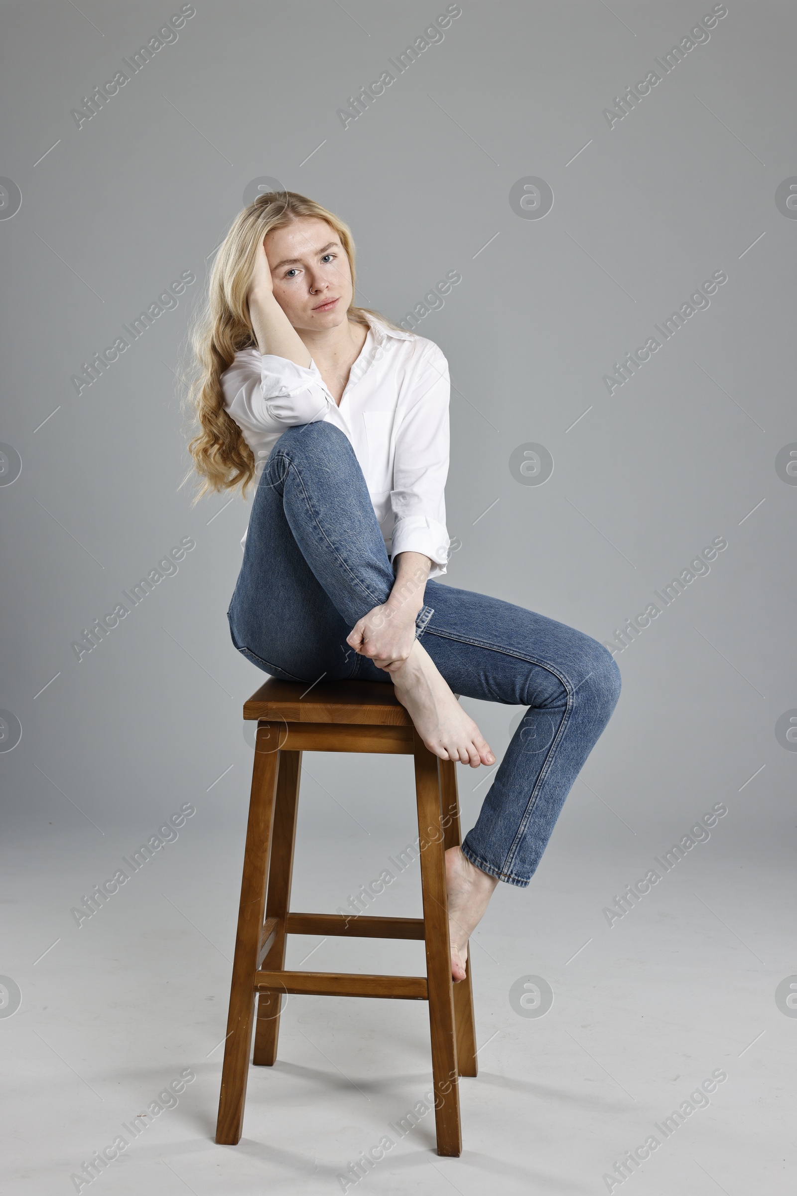 Photo of Beautiful young woman in stylish jeans sitting on stool against grey background