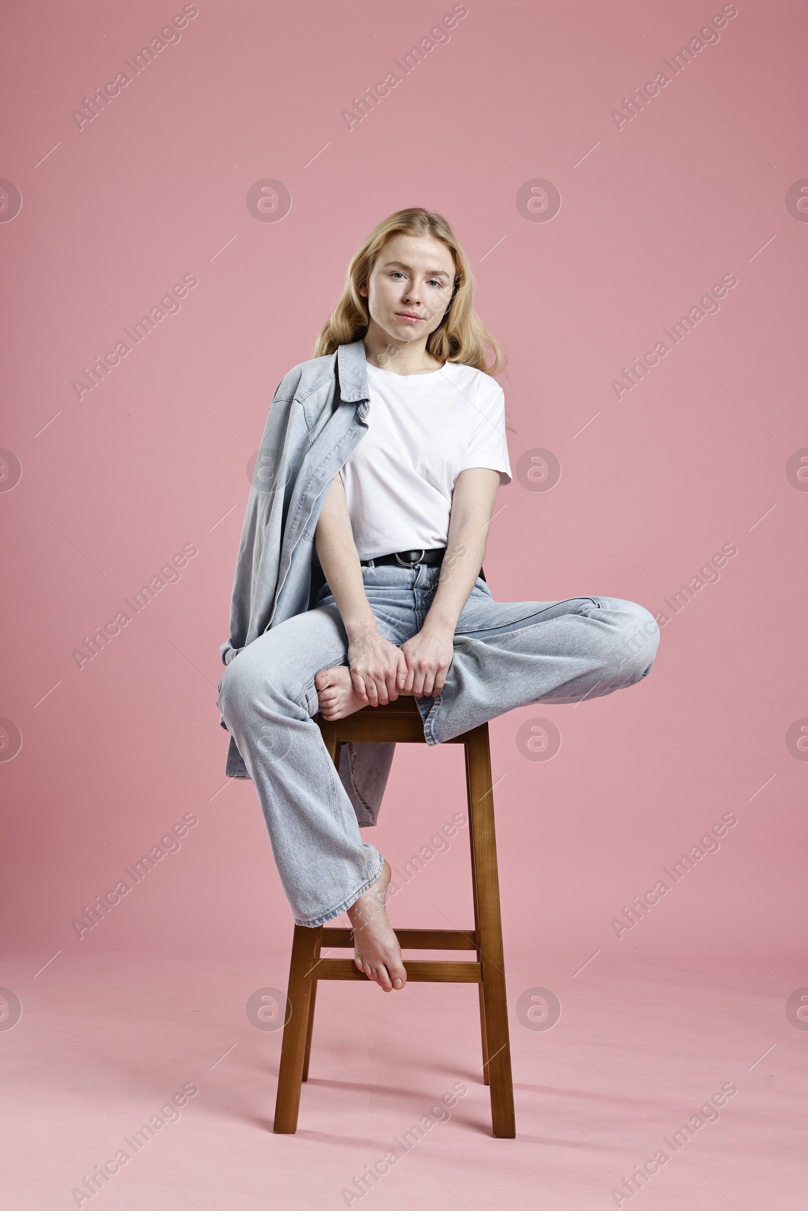 Photo of Beautiful young woman in stylish jeans sitting on stool against pink background