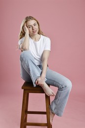 Smiling woman in stylish jeans sitting on stool against pink background