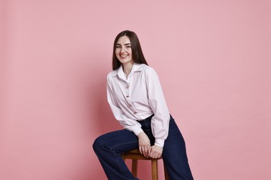 Photo of Smiling woman in stylish jeans sitting on stool against pink background