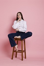 Photo of Beautiful young woman in stylish jeans sitting on stool against pink background