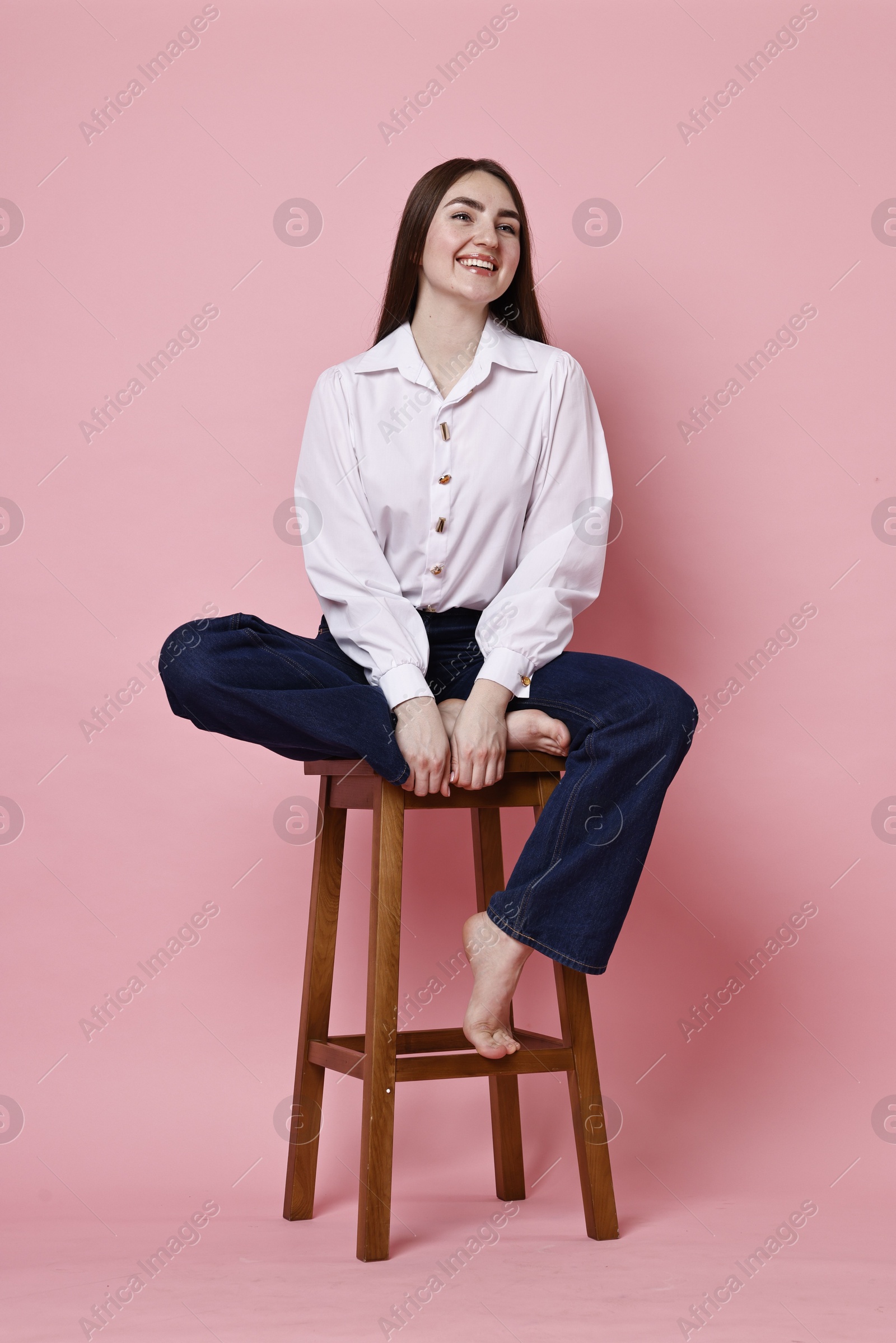 Photo of Smiling woman in stylish jeans sitting on stool against pink background