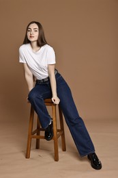 Photo of Beautiful young woman in stylish jeans sitting on stool against brown background