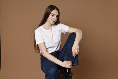 Photo of Beautiful young woman in stylish jeans sitting on stool against brown background