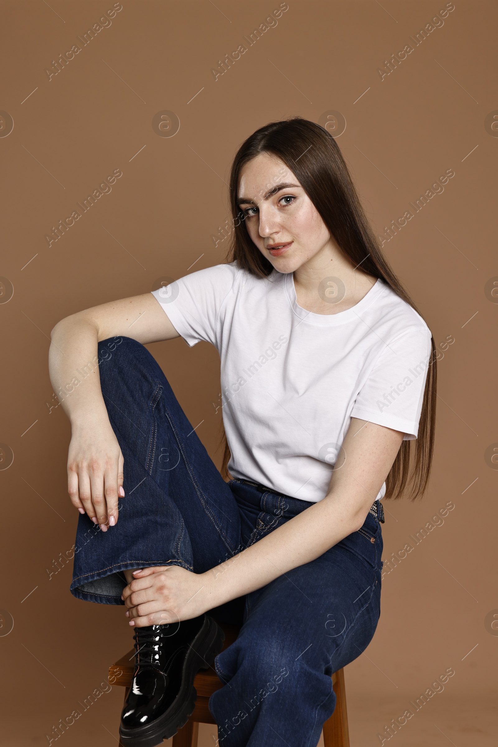 Photo of Beautiful young woman in stylish jeans sitting on stool against brown background