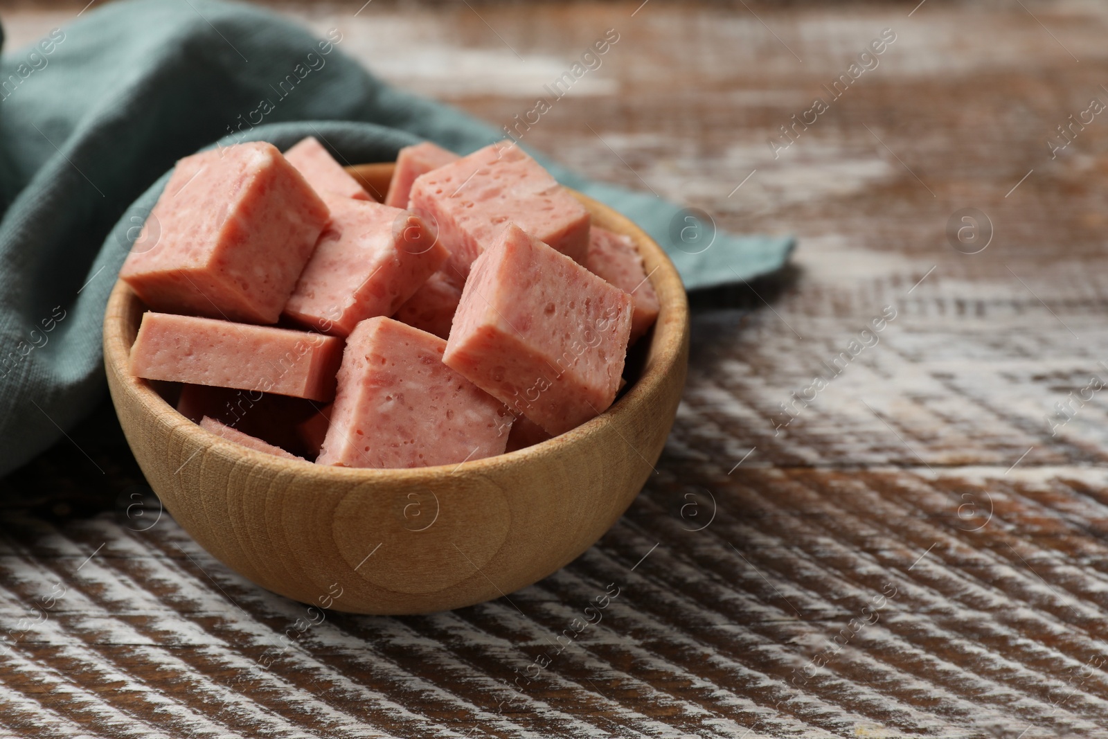 Photo of Pieces of canned meat in bowl and towel on wooden table, closeup. Space for text