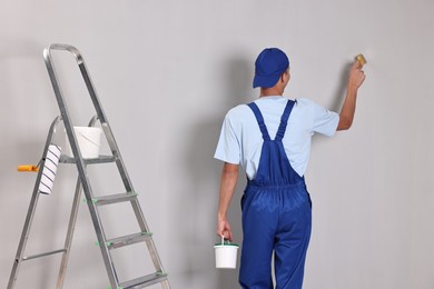 Photo of Male handyman painting wall with brush near ladder indoors, back view