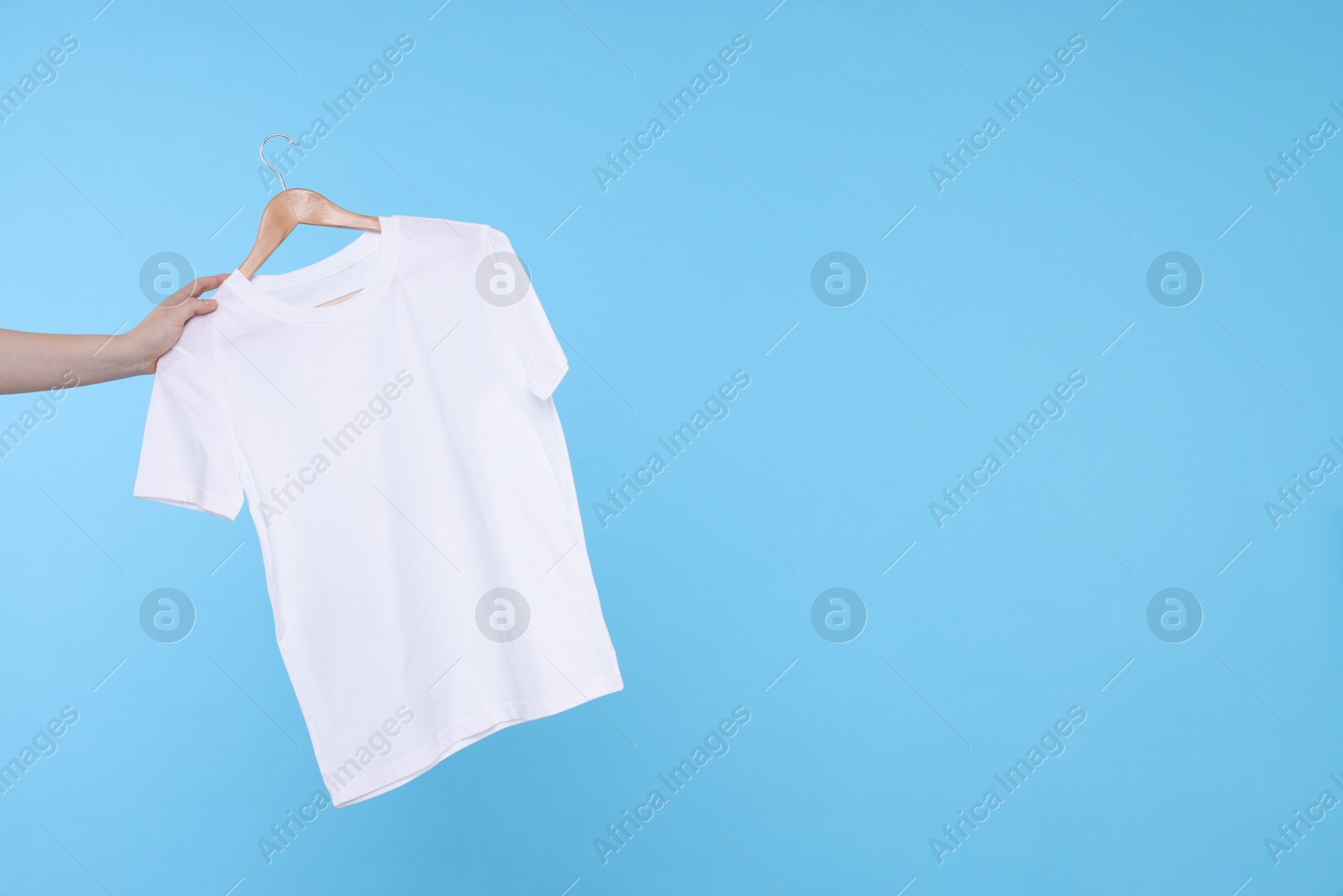 Photo of Woman holding hanger with white t-shirt on light blue background, closeup. Space for text