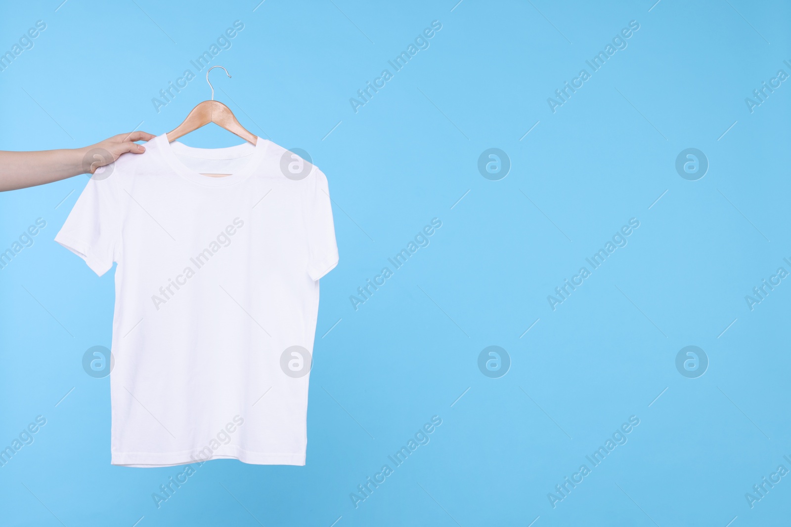 Photo of Woman holding hanger with white t-shirt on light blue background, closeup. Space for text