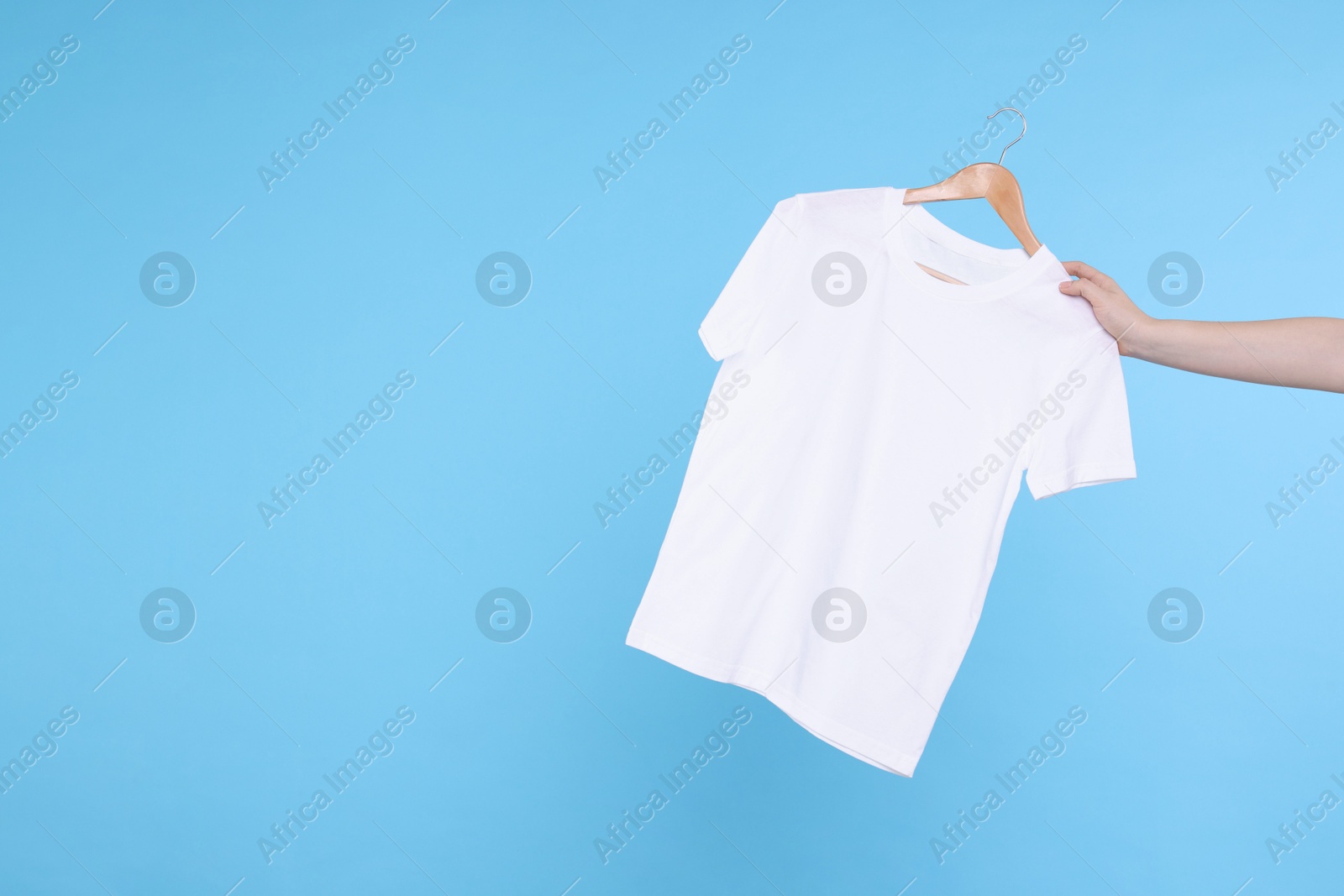 Photo of Woman holding hanger with white t-shirt on light blue background, closeup. Space for text